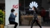 A woman wearing a face mask to help curb the spread of the coronavirus browses her smartphone as a masked woman walks by the Huawei retail shop promoting it 5G network in Beijing Oct. 11, 2020.