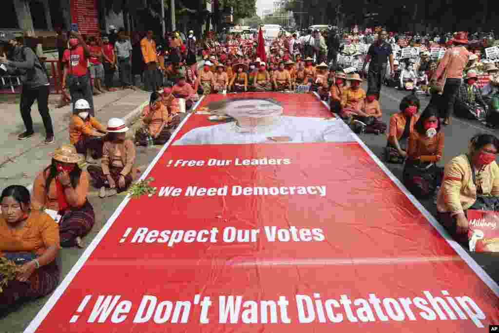 Protesters sit next to a giant banner with images of detained leader Aung San Suu Kyi during an anti-coup protest in Mandalay, Myanmar.